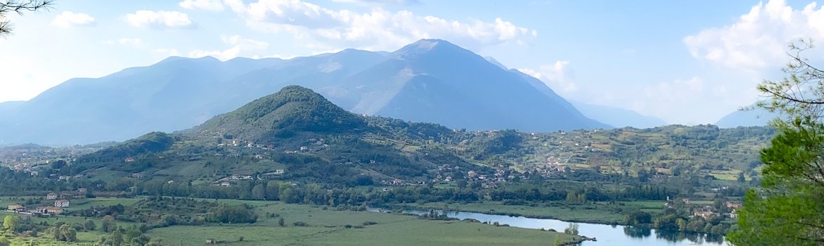A green landscape. Middle and background: mountains. A river meanders in the foreground.